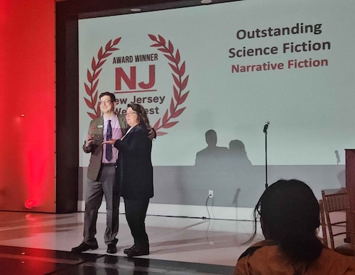 left to right: Voice actor AJ Fidalgo and writer Lindsay Harris Friel accept the award for Outstanding Science Fiction on stage at New Jersey Web Fest 2026.